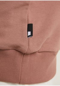 Sudadera rosa con ribetes en el bajo y los puños, textura suave y una pequeña etiqueta de logotipo en blanco y negro en el costado.