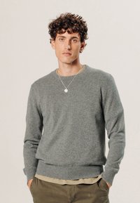 Grauer Wollpullover mit Rundhalsausschnitt, gerippten Bündchen und Saum, über einem beigen Hemd getragen. Bietet eine dezente Textur und eine lockere Passform.