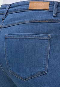 Vue de dos en gros plan d'un jean en denim bleu avec une seule poche et une étiquette de marque en cuir beige inscrite ONLY à la ceinture.