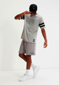 T-shirt gris con rayas negras, con el logo de "ELLESSE", combinado con pantalones cortos grises y zapatillas blancas. El modelo lleva una gorra negra.