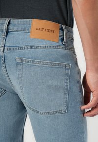Lyseblå denimjeans med fem lommer, som har et brun skinnlapp merket "ONLY & SONS" på baklivet.