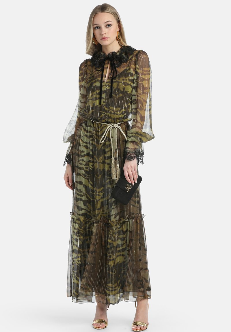 Frau in einem langen, transparenten Kleid mit Tiermuster und schwarzem Spitzenkragen, mit Quasten-Gürtel, goldenen Sandalen, goldenen Creolen und einer schwarzen Clutch-Tasche.