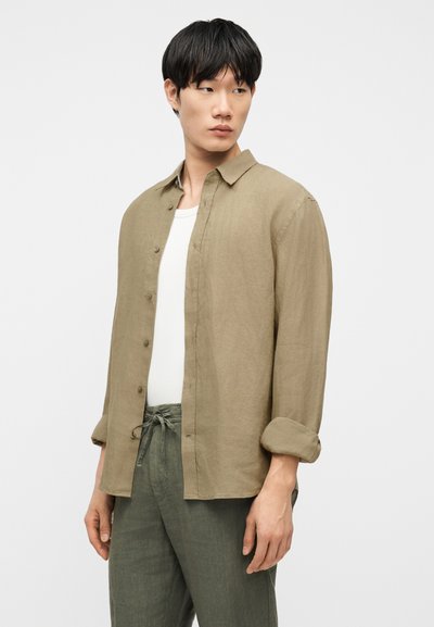 Νέος άνδρας που φοράει ανοιχτό μπεζ πουκάμισο με κουμπιά πάνω από λευκό τοπ και χακί παντελόνι με κορδόνι, κοιτάζοντας πλάγια σε απλό φόντο.