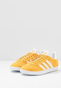 Gula mockaskor med vita ränder och accenter, som har en klassisk design och strukturerad yta. Modellenamn "Gazelle" på sidan.