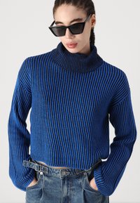 Blå og svart stripete strikkegenser med høy krage og løs passform. Brukt med høyt liv jeans.