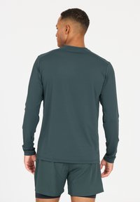 Langärmliges Sportshirt in dunklem Teal, aus feuchtigkeitsableitendem Stoff mit glatter Textur; ausgestattet mit Flachnähten für zusätzlichen Komfort.