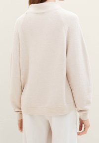 Pull rose pâle avec un col montant, une texture côtelée et une coupe décontractée. Présente des épaules tombantes et des bords de manches retroussés. Design minimaliste.