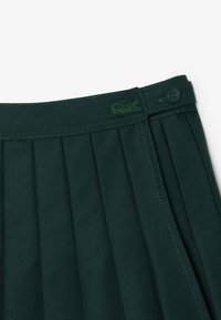 Jupe plissée verte en tissu texturé, avec un logo brodé à la taille et un détail de couture verte contrastante.