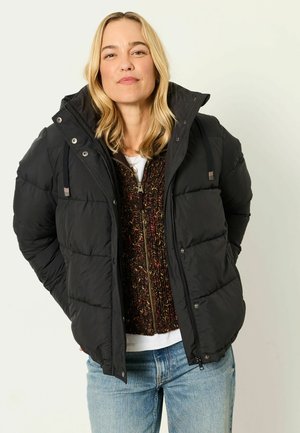 Donna con giacca imbottita nera sopra un maglione lavorato a maglia multicolore e jeans blu, in piedi davanti a uno sfondo chiaro semplice.