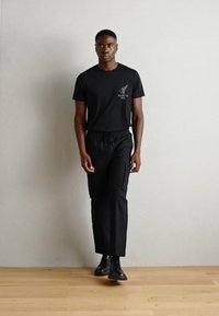 Black t-shirt with logo, paired with black pants featuring pockets. Model stands on wooden floor against a light gray wall, wearing black boots.