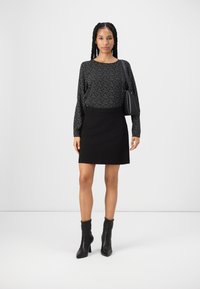 Blouse noire à manches longues avec des taches blanches, associée à une jupe noire. Le mannequin porte des bottes à talons noires et une petite sacoche noire.