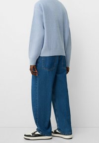 Person, die einen hellblauen Strickpullover, weite blaue Jeans und schwarz-weiße Turnschuhe trägt, vor einem schlichten weißen Hintergrund steht.