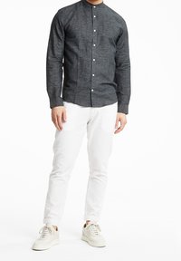 Ciemnoszara koszula z długim rękawem i kołnierzykiem mandarynki, z przodu zapinana na guziki, zestawiona z białymi spodniami slim-fit i jasnymi trampkami.