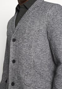 Grå tekstureret blazer med reverskrave, sorte knapper og en enkelt brystlomme, lagdelt over en mørk skjorte.