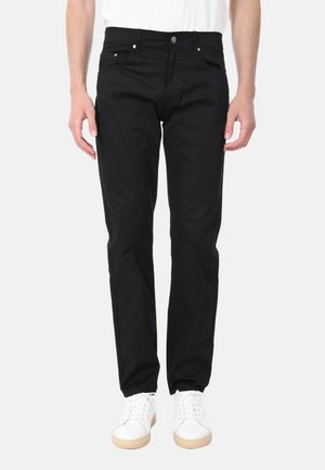 Uomo con jeans neri dritti e sneakers bianche in piedi contro uno sfondo chiaro semplice, mani rilassate lungo i fianchi.