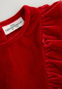 Czerwony aksamitny top z okrągłym dekoltem i falbaniastym akcentem z boku. Metka z widoczną informacją o rozmiarze i napis „Coccodrillo”.