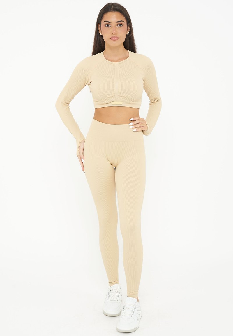 Set di activewear beige a coste composto da un top corto a maniche lunghe e leggings a vita alta, realizzato in materiale elastico. Le sneakers bianche completano il look.