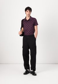 Vijolična polo majica s vzorcem, črne cargo hlače in črni loafers. Model drži črno torbo in stoji proti belemu ozadju.