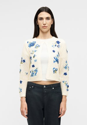 Kvinde iført en cremet strikket cardigan med blå blomstermotivbroderi over en hvid top og mørke jeans, stående foran en ensfarvet baggrund.