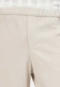 Beige sportieve broek met een elastische tailleband, voorzien van een zijzak en een gladde, lichte stoftextuur.