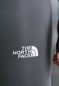 Tummanharmaa kangas, jossa valkoinen The North Face -logo ja tumman sävyinen käsi pitää kankaan reunaa oikealla puolella.