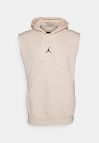 Sleeveless hoodie in light beige with a front pocket, drawstring hood, and a black Jumpman logo on the chest. Soft cotton blend material.