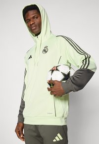 Lys grønn hettegenser med svarte detaljer, med glidelås, Real Madrid-logo, og tre svarte striper på ermene, sammen med en fotball.