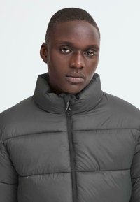 Grå pufferjakke med høj krave, quiltet design og frontlynlås. Materialet ser glat og isoleret ud, velegnet til koldt vejr.