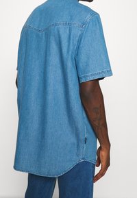 Světle modrá denimová košile s krátkým rukávem, zaobleným lemem a detailem na yoke vzadu. Disponuje ležérním prošíváním a volným střihem.