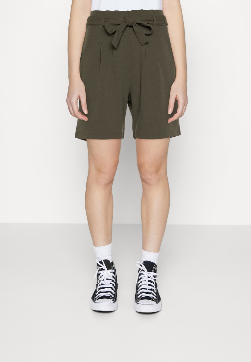 Shorts taille haute vert olive avec une ceinture nouée, des plis à l'avant et une coupe décontractée. Associés à des baskets montantes noires et des chaussettes blanches.