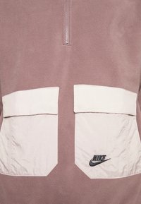 Un haut en polaire lilas clair avec un demi-zip, doté de deux poches rectangulaires crème et d'un logo Nike noir sur la poche inférieure gauche.