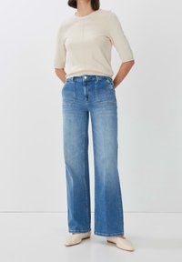 Vaaleanbeige lyhythihainen paita, jossa on pyöreä kaula-aukko, yhdistettynä korkeavyötäröisiin, vaaleansinisiin levyjenlahkeisiin farkkuihin, joissa on etutaskut ja nappikiinnitys.