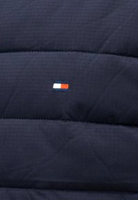 Ναυτικό μπλε καπιτονέ ύφασμα με μικρό ορθογώνιο λογότυπο που διαθέτει λευκά, κόκκινα και ναυτικά μπλε χρωματικά μπλοκ ραμμένα στο κέντρο.