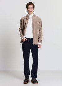 Beige Hemd zum Knöpfen über einem weißen Rollkragenpullover, kombiniert mit dunkelblauen Hosen und braunen Schuhen. Glatter Stoff, lockere Passform und dezente Texturen.