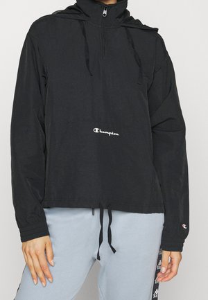 Osoba ubrana w czarną bluzę Champion z kapturem, z logo z przodu, półzamkiem, ściągaczami oraz jasnoszare spodnie dresowe z czarnymi paskami po bokach.