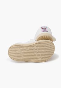 Zapato infantil de tela blanca con una suave suela de goma beige. Presenta una etiqueta bordada colorida que representa a un mono y el texto: "Hecho en España."