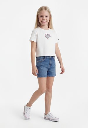 Ung jente smiler, iført en hvit cropped t-skjorte fra Tommy Hilfiger, denimshorts og hvite sneakers, står på ensfarget hvit bakgrunn.