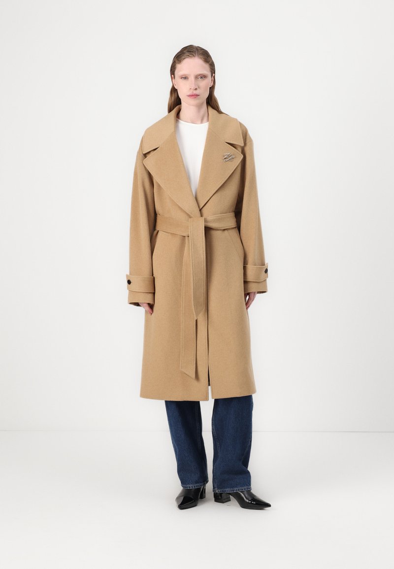 Cappotto trench in lana beige con vita abbeltata, colletto ampio e polsini abbottonati. Presenta una spilla metallica sulla fodera. Indossato su pantaloni in denim.