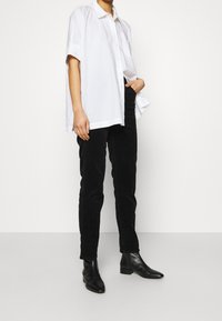 Bílé nadměrné krátké rukávové košilové tričko v kombinaci s těsnými černými manšestrovými kalhotami a černými koženými kotníkovými botami na podpatku. Hladké textury tkanin.