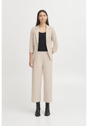 Femme debout, mains détendues, portant un blazer beige et un pantalon large, un haut noir et des bottines noires sur fond blanc.