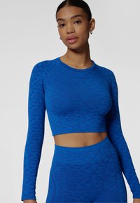 Niebieski sportowy krótki top z długimi rękawami, teksturowanym wzorem i detalem z logo. Materiał wydaje się elastyczny i dopasowany.