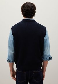 Gilet lavorato a maglia blu navy con texture a coste, indossato sopra una camicia di jeans azzurro chiaro, con le maniche arrotolate e jeans scuri su misura.