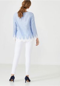 Ljusblå långärmad blus med vit scallop-lace trim vid nederkanten, parat med vita slim-fit byxor och mörkblå högklackade skor.