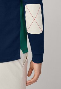 Marineblaues Langarmhemd mit grünem seitlichem Einsatz und weißen Ellbogenpatch, der mit rotem Z stitching verziert ist. Weicher Stoff, figurbetontes Design und kontrastierende Farben.