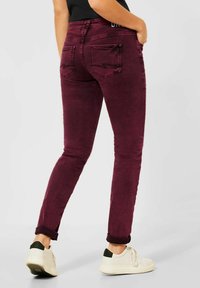 Jeans skinny burdeos oscuro en tela de denim, con un diseño clásico de cinco bolsillos y dobladillos enrollados, combinados con zapatillas blancas.