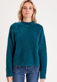 Suéter texturizado en color verde azulado con cuello alto y mangas largas, con un corte holgado. Combinado con jeans de mezclilla azul oscuro.