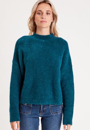 Maglioncino teal con texture, col alto e maniche lunghe, caratterizzato da una vestibilità ampia. Abbinato a jeans in denim blu scuro.