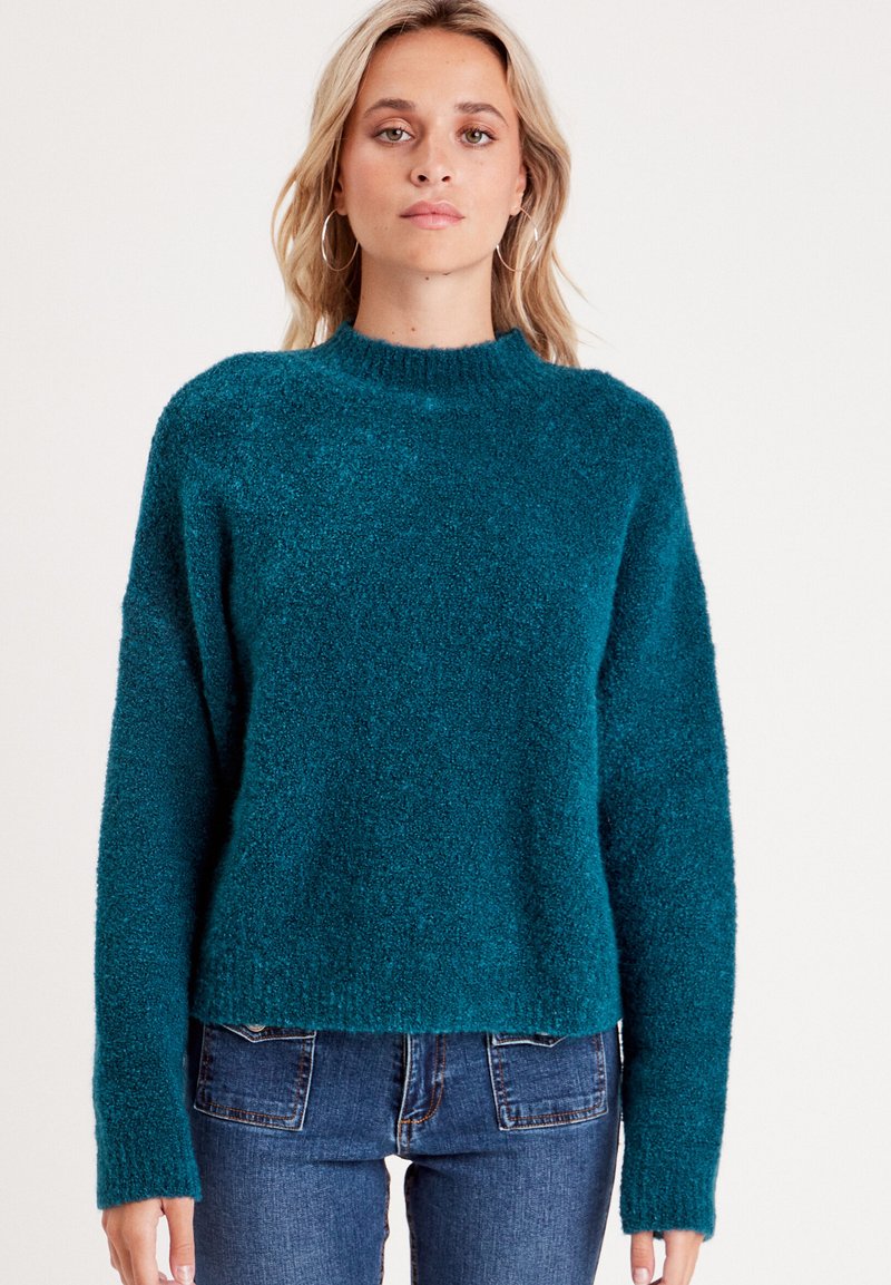 Suéter texturizado en color verde azulado con cuello alto y mangas largas, con un corte holgado. Combinado con jeans de mezclilla azul oscuro.