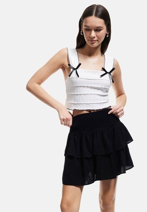 Hvid cropped top med teksturerede striber og sorte sløjfe-accenter, parret med en sort volangskørt med smocket talje.