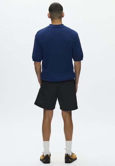 Polo bleu à manches courtes avec texture en forme de losange, porté avec un short noir. Le mannequin porte des chaussures jaunes et noires avec des chaussettes blanches, vu de dos.